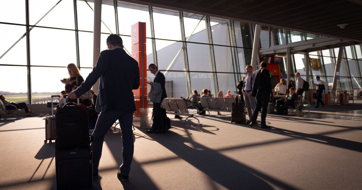 A traveller pulling wheeled suitcases past airport departure gate seating, backlit by low evening sun