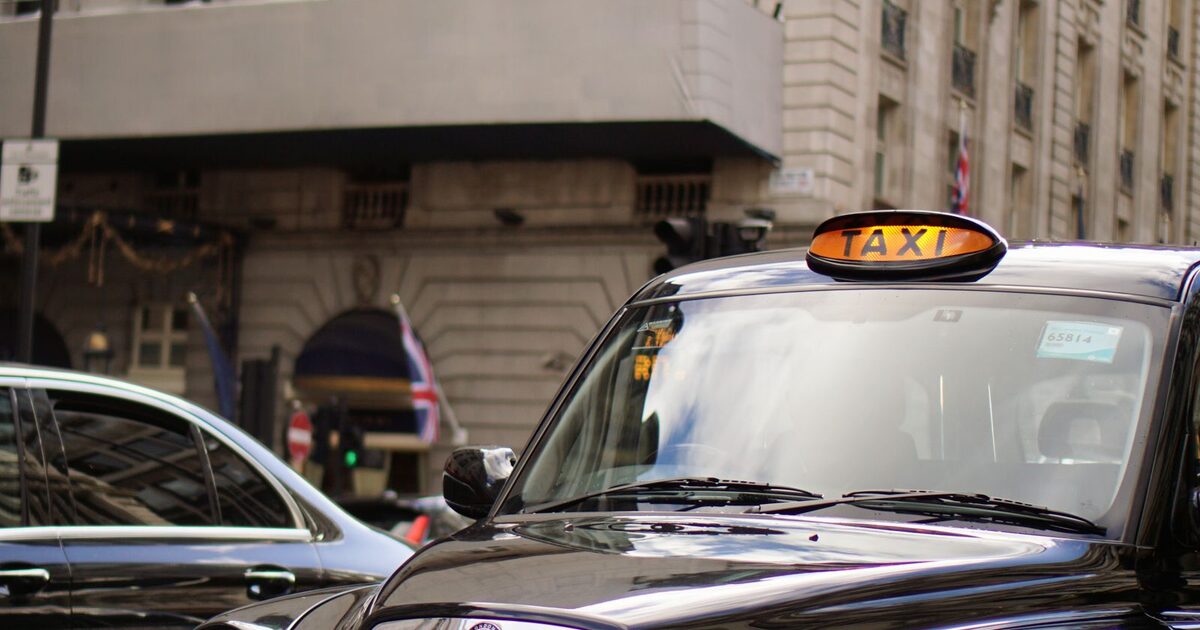 A traditional black London taxi with its illuminated TAXI sign, parked on a street lined with period architecture